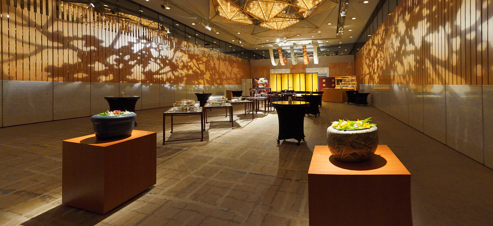 広々としたイベントホールの全体像。木目調の壁の前面には、影絵のような木々のシルエットが壁の大部分に映し出されている。部屋の中央にはいくつものテーブルと椅子が配置され、手前には装飾された植物の鉢が2つ置かれている。