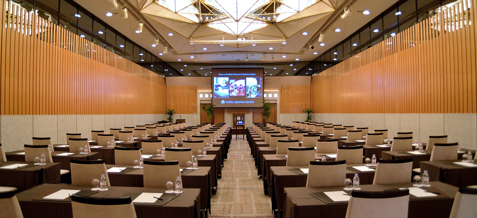 広々とした会議室の全体像です。部屋の中央には、多数の会議用テーブルと椅子が整然と横一列に並べられています。部屋の奥には大きなスクリーンが設置されており、その下には演台のような台が見えます。壁は木目調で、天井は複数のパネルで構成され、照明が備え付けられています。