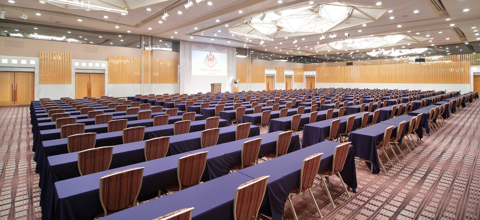 広々としたイベントホールの全体像です。手前から奥に向かって、非常に多くの長机と椅子が整然と並べられています。机には紺色のクロスがかかっており、椅子は木製の背もたれと座面が特徴です。部屋の奥には、白いスクリーンが設置されており、そのスクリーンには赤い模様と文字が映し出されています。部屋の壁は木目調で、上部には複数の照明が配置されています。