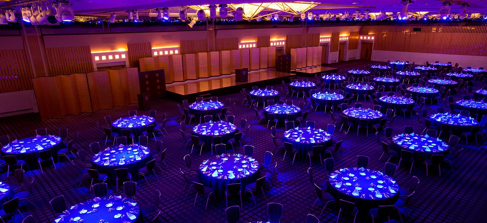 華やかな照明で彩られた広々としたイベントホールの全体像です。室内は主に紫色と青色のライトで照らされ、幻想的な雰囲気を醸し出しています。手前から奥にかけて、多数の丸いテーブルが配置されており、それぞれのテーブルには青い照明が当てられ、テーブルの表面には白い模様が浮かび上がっています。