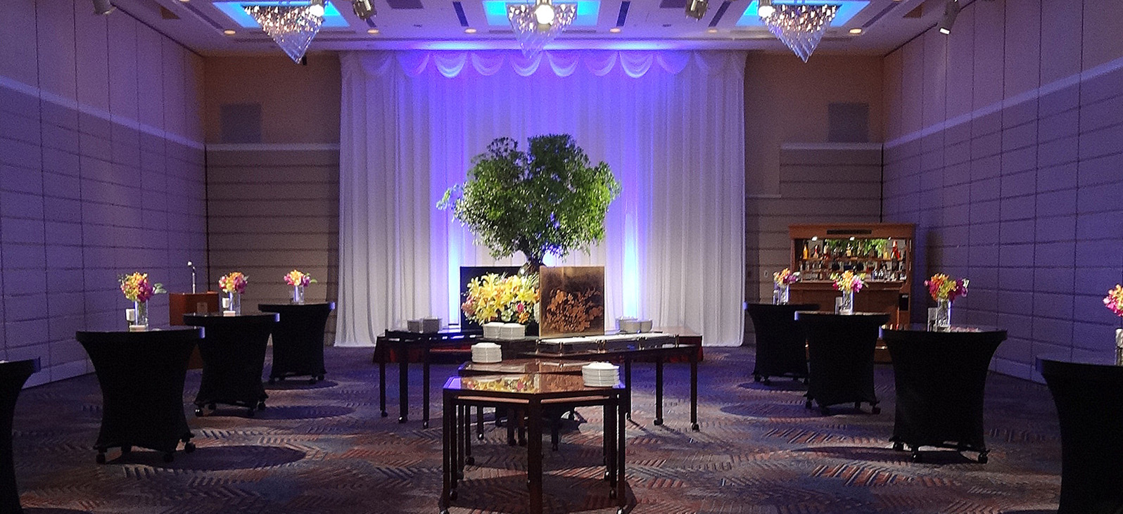 白い幕が青いライトで照らされ、中央に大きな盆栽が飾られたイベント会場。会場には黒い布で覆われた円卓や、木製のテーブルが複数配置され、カラフルな花のアレンジメントが飾られている。右奥にはアルコール飲料が並んだバーカウンターが見える。