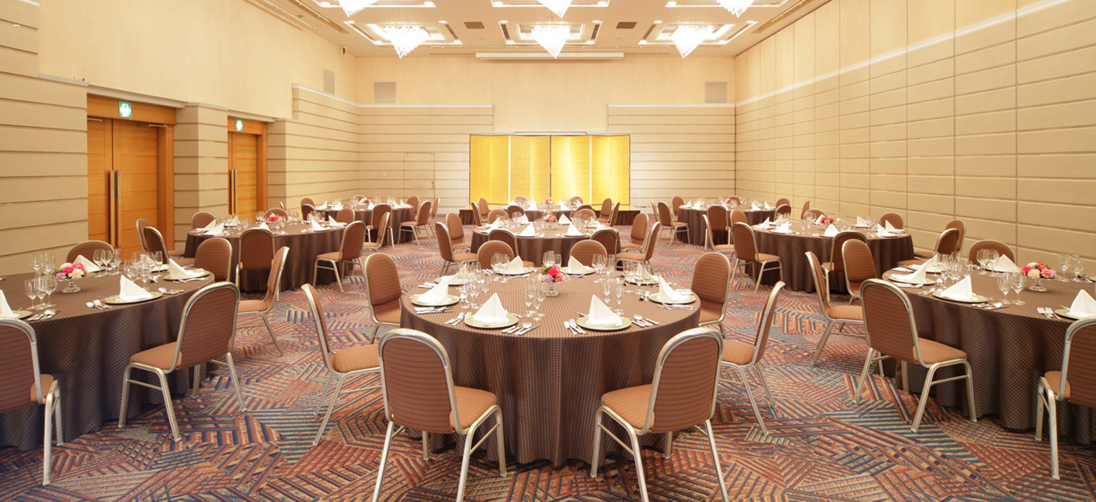 複数の丸テーブルと椅子が整然と並べられた、広々とした宴会場。各テーブルには白い食器とグラス、ピンク色のナプキンがセッティングされ、小さな花のアレンジメントが飾られている。会場の壁はベージュ色で、天井にはシャンデリアとダウンライトが多数設置されている。