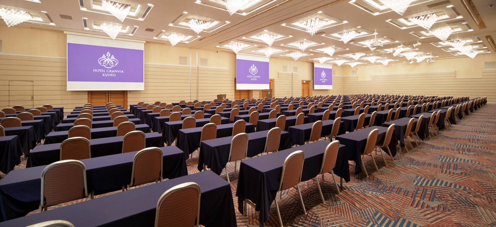 天井に多数のシャンデリアとダウンライトが輝く、広々とした会議室。会場には、紺色のクロスがかけられた長テーブルと茶色の椅子が、講演会形式で整然と並べられている。部屋の奥には3つの大きなスクリーンが設置されており、それぞれに「HOTEL GRANVIA KYOTO」のロゴが表示されている。