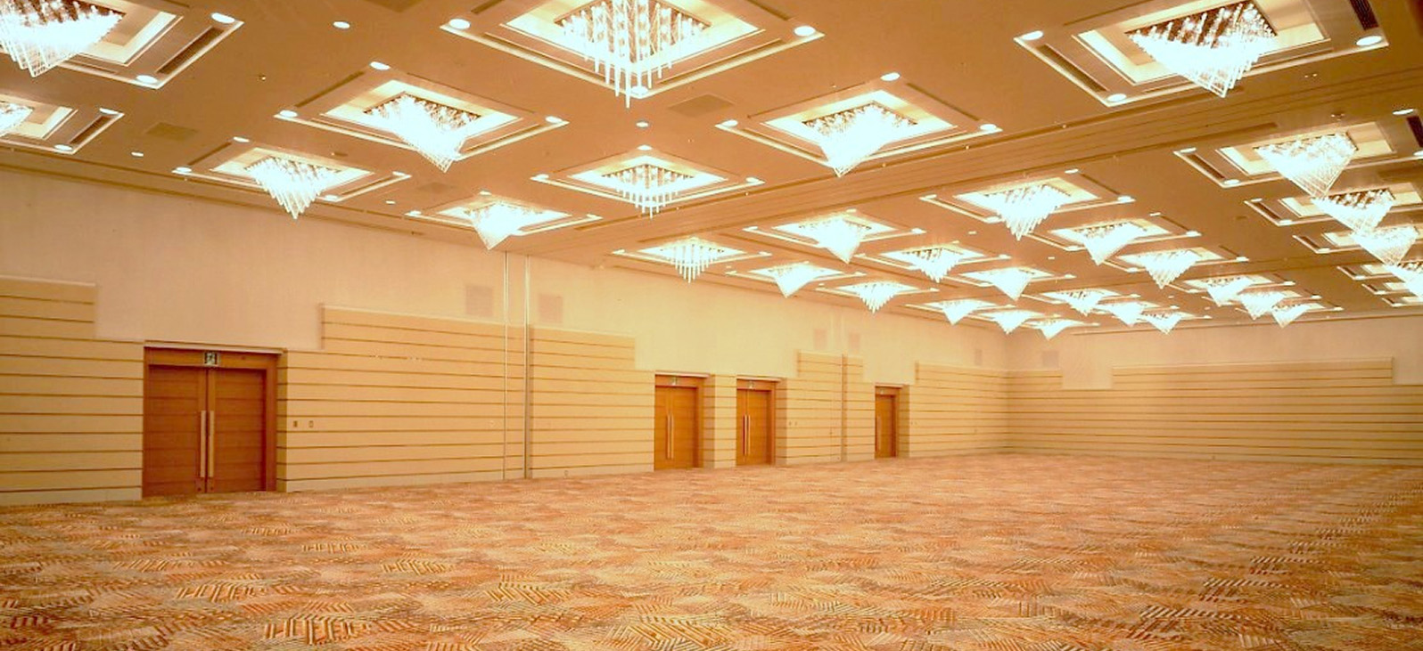 華やかなシャンデリアと多数のダウンライトで照らされた、広々とした宴会場の様子。ベージュの壁には木製の扉がいくつか並び、床にはオレンジとベージュの幾何学模様のカーペットが敷かれている。