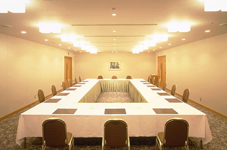 ベージュの壁と明るい照明の、コの字型（U字型）に会議テーブルが配置された中規模な会議室