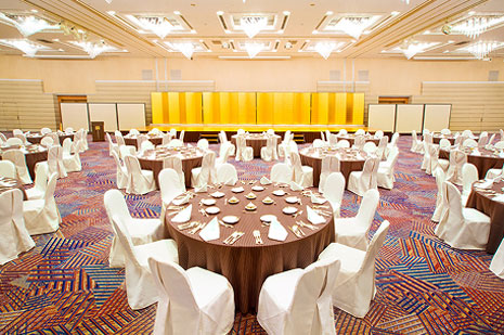 明るい照明の、白い椅子とクリーム色のテーブルクロスがかけられた丸テーブルが多数配置された大規模な宴会場