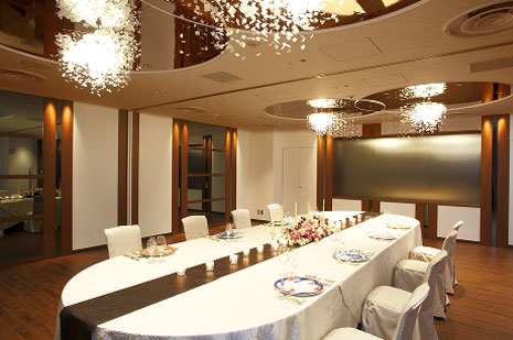 茶色を基調とした内装の、白いテーブルクロスがかけられた長テーブルが配置された役員会議室または小宴会場