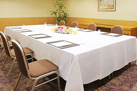明るい照明の、白いテーブルクロスがかけられた長テーブルが配置され、花が飾られた小会議室