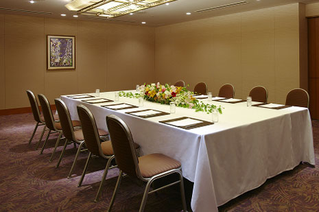 明るい照明の、白いテーブルクロスがかけられた長テーブルが配置され、椅子が並んだ小会議室
