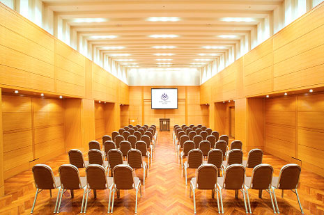 木目調の壁と明るい照明の、ステージに向かってシアター形式で椅子が並べられた中規模な会議室
