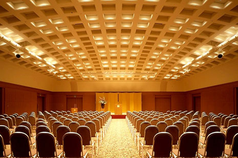 高い天井と木目調の壁、ステージに向かってシアター形式で多数の椅子が並べられた会議室または宴会場