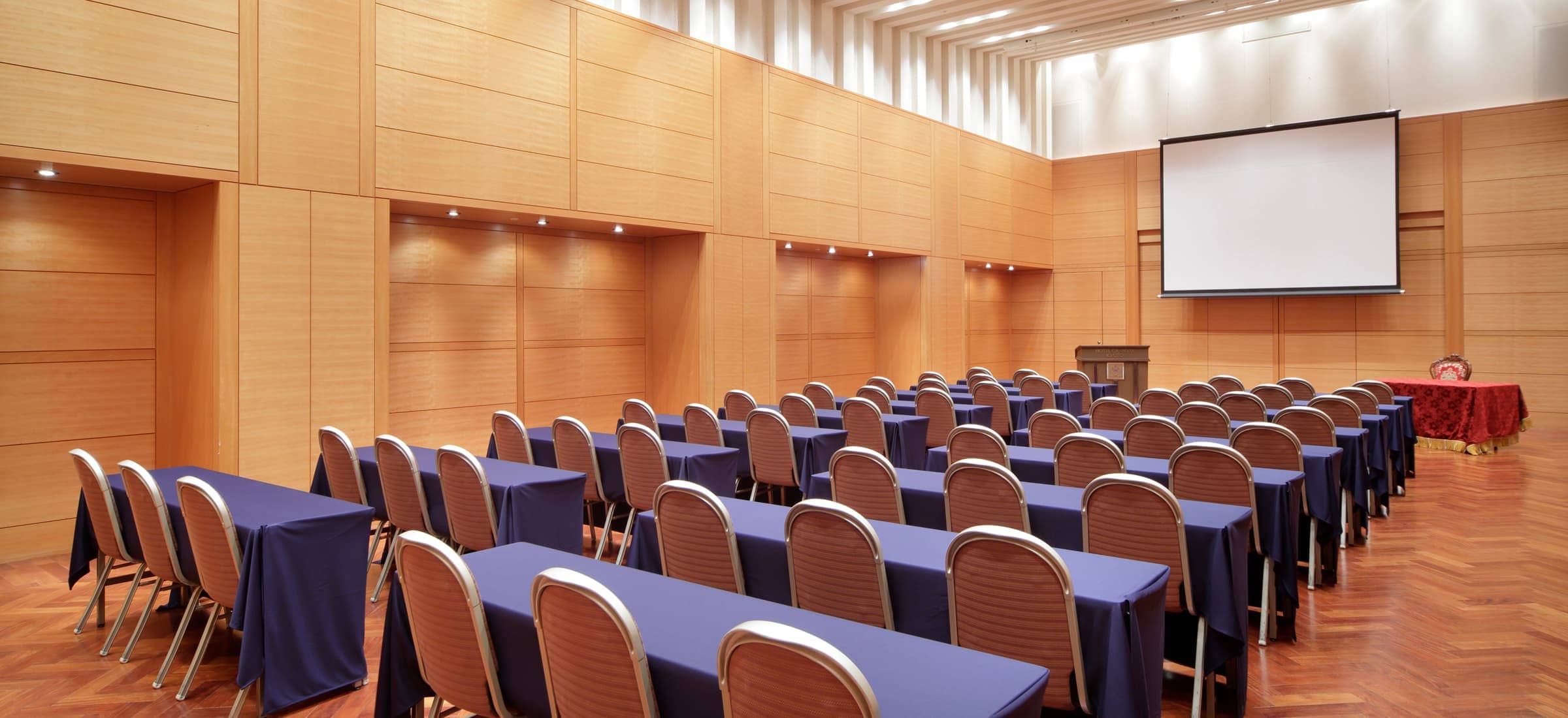 広々としたイベントホールの全体像。木目調の壁の前面には、影絵のような木々のシルエットが壁の大部分に映し出されている。部屋の中央にはいくつものテーブルと椅子が配置され、手前には装飾された植物の鉢が2つ置かれている。