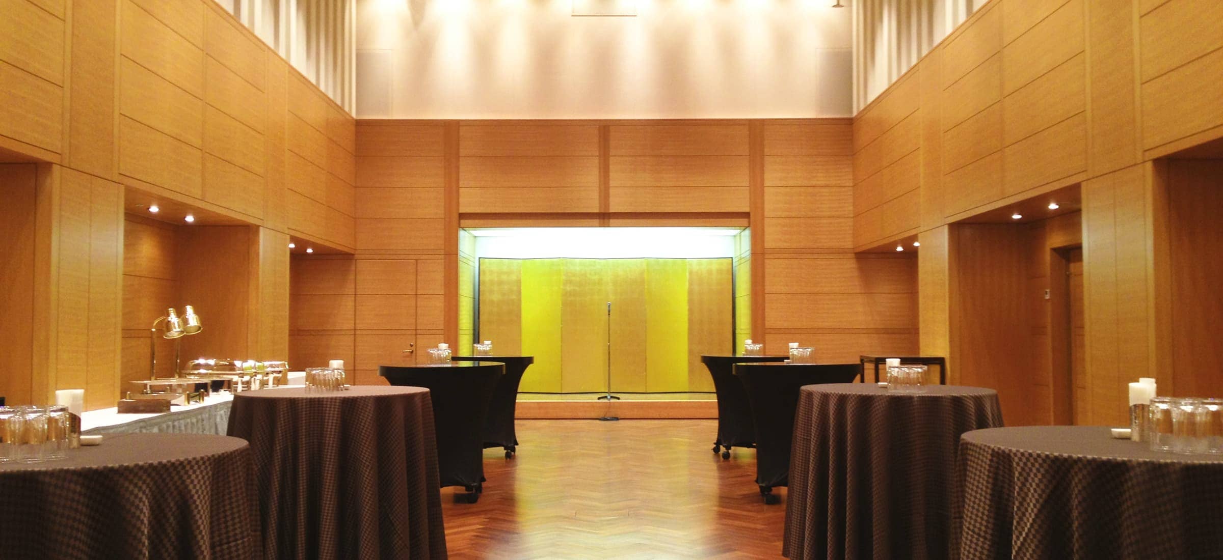 広々としたイベントホールの全体像です。手前から奥に向かって、非常に多くの長机と椅子が整然と並べられています。机には紺色のクロスがかかっており、椅子は木製の背もたれと座面が特徴です。部屋の奥には、白いスクリーンが設置されており、そのスクリーンには赤い模様と文字が映し出されています。部屋の壁は木目調で、上部には複数の照明が配置されています。