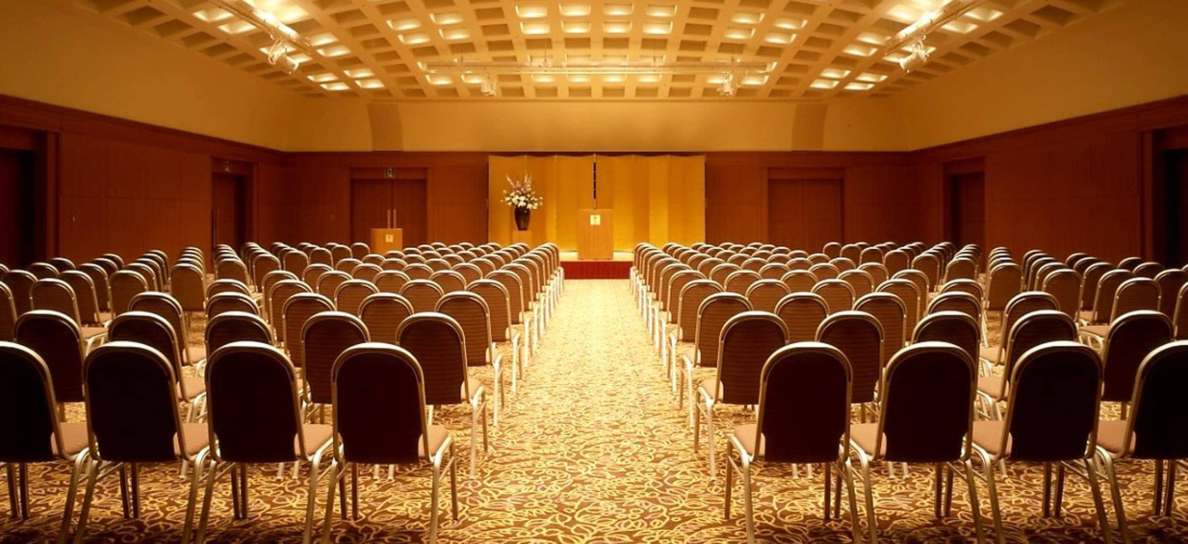 広々としたイベントホールの全体像。木目調の壁の前面には、影絵のような木々のシルエットが壁の大部分に映し出されている。部屋の中央にはいくつものテーブルと椅子が配置され、手前には装飾された植物の鉢が2つ置かれている。