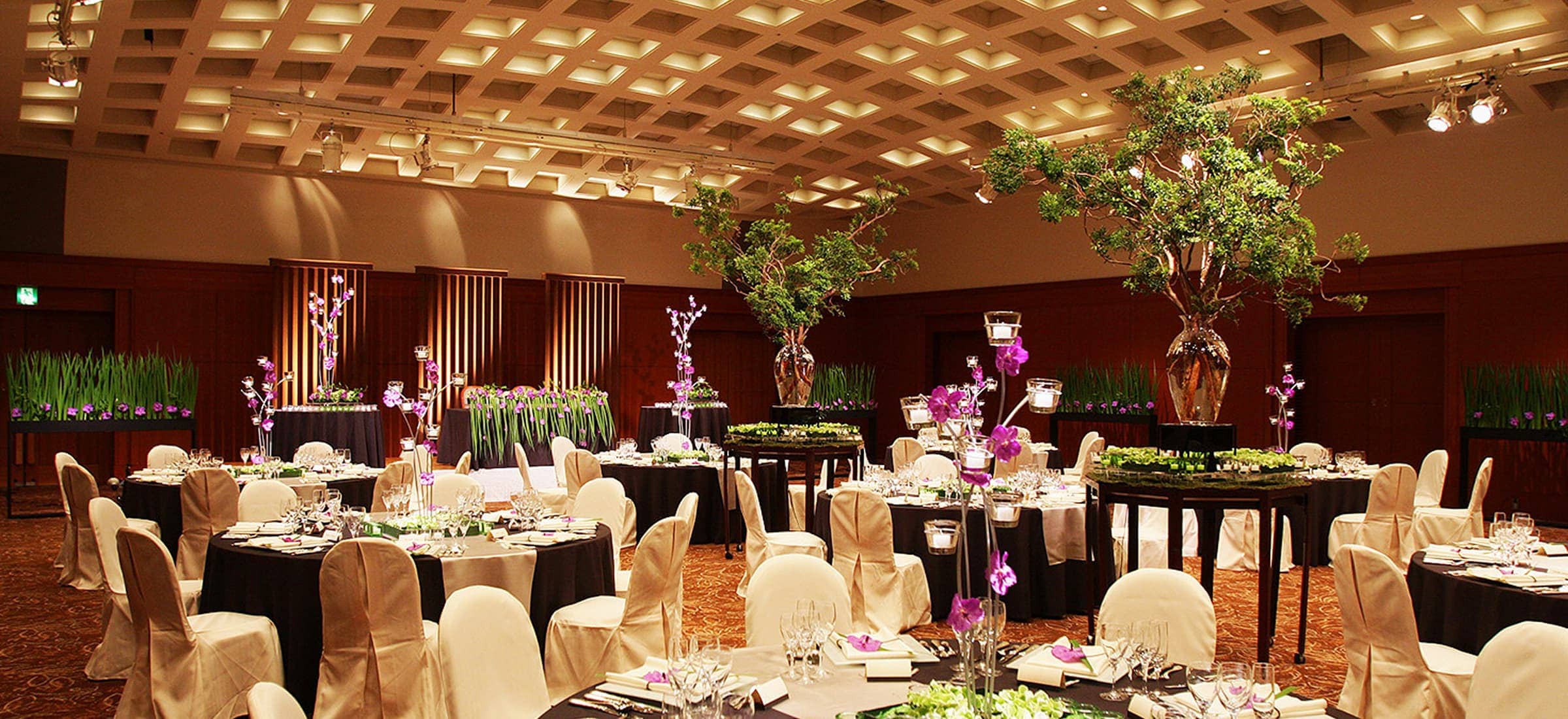 広々とした会議室の全体像です。部屋の中央には、多数の会議用テーブルと椅子が整然と横一列に並べられています。部屋の奥には大きなスクリーンが設置されており、その下には演台のような台が見えます。壁は木目調で、天井は複数のパネルで構成され、照明が備え付けられています。