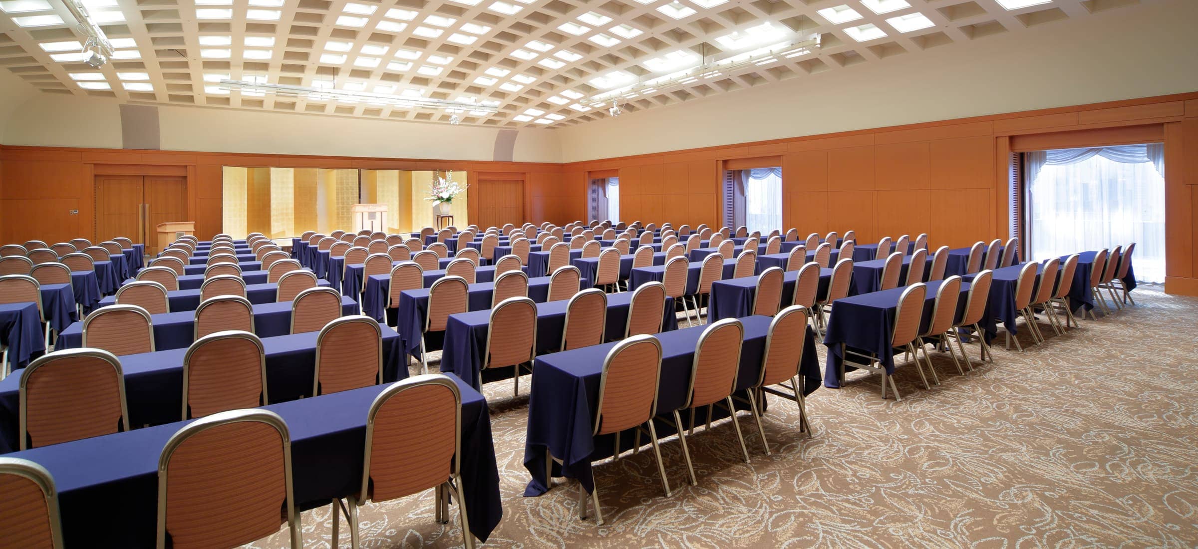 広々としたイベントホールの全体像です。手前から奥に向かって、非常に多くの長机と椅子が整然と並べられています。机には紺色のクロスがかかっており、椅子は木製の背もたれと座面が特徴です。部屋の奥には、白いスクリーンが設置されており、そのスクリーンには赤い模様と文字が映し出されています。部屋の壁は木目調で、上部には複数の照明が配置されています。