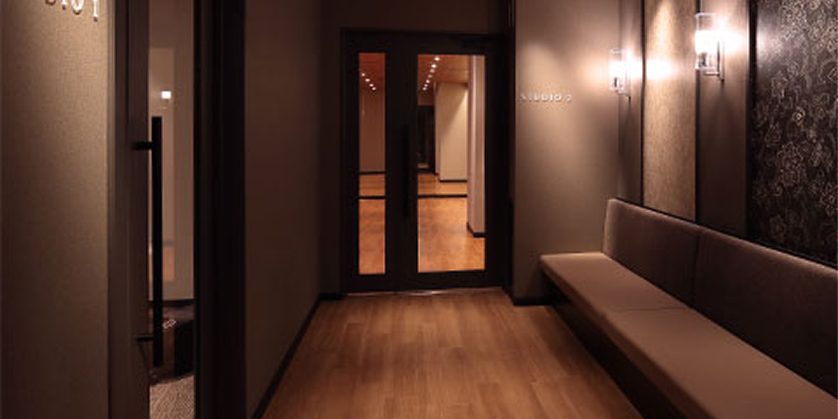 グループレッスンスタジオ前の待合廊下。長いソファベンチと落ち着いた照明が、リラックスできる空間を演出している。