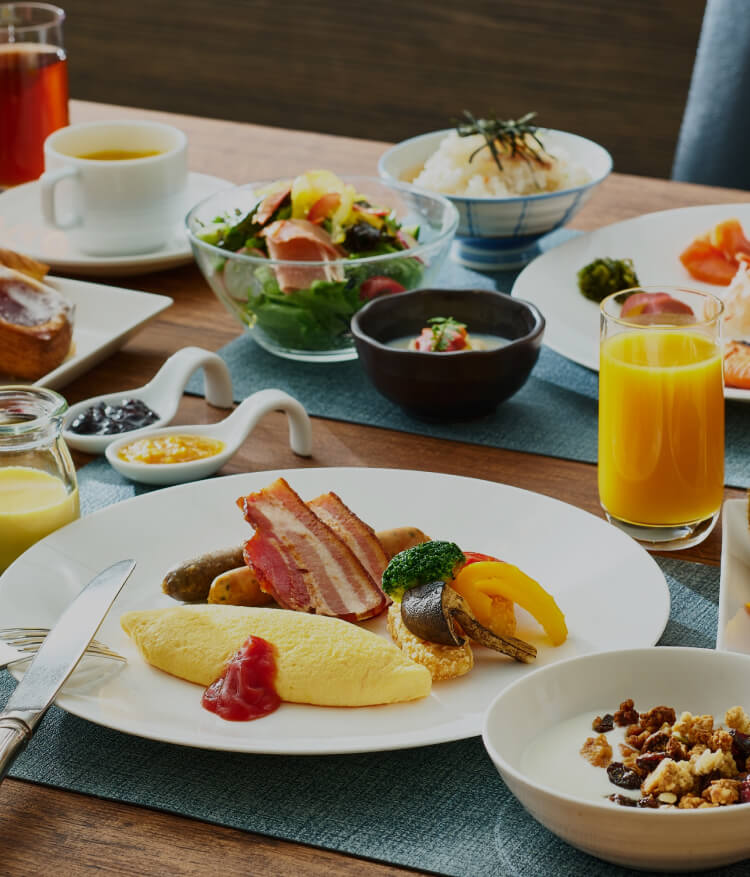焼きたてのパン、オムレツとベーコン、サラダ、和食（ご飯と味噌汁）、カットフルーツ、シリアル、ジュース、コーヒーなど、多彩なメニューが並ぶ豪華な朝食のテーブル。