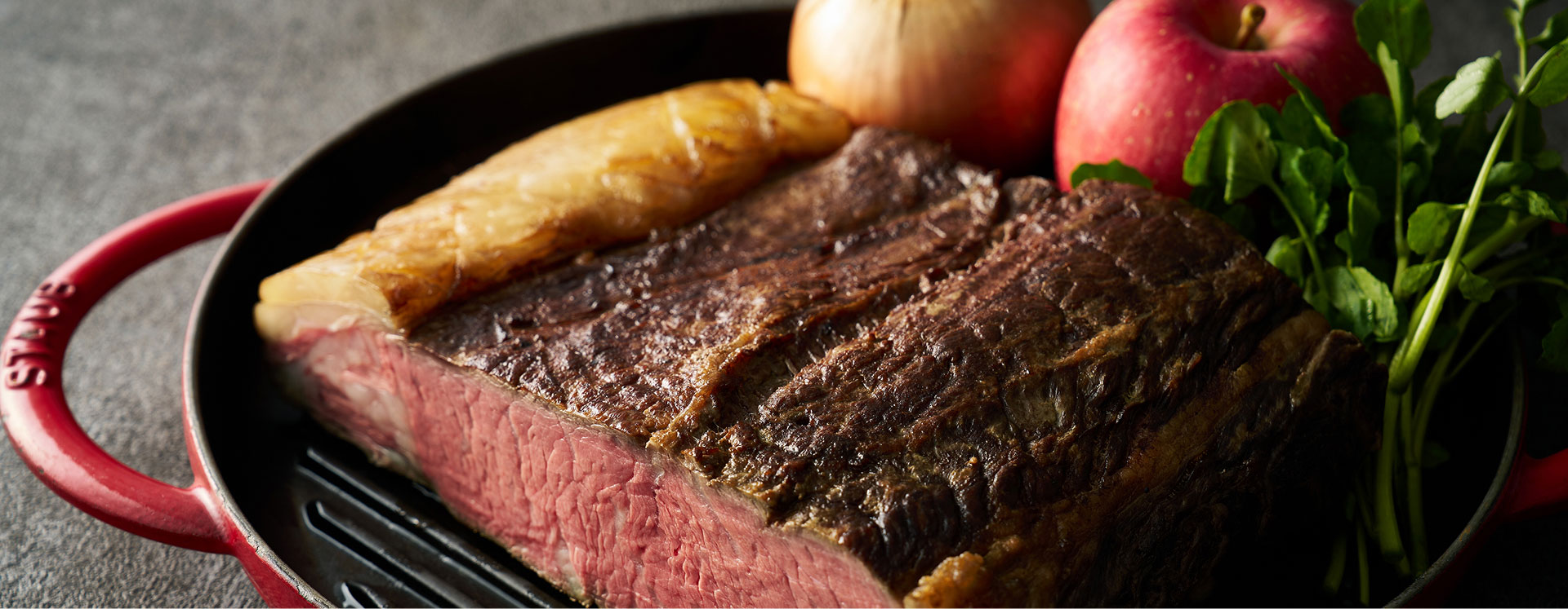 鉄鍋に盛られたローストビーフ。断面が美しく焼かれ、リンゴや野菜が添えられている。