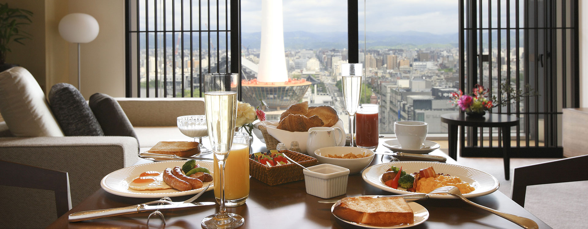 大きな窓のそばで楽しむ朝食。ジュース、パン、卵料理などが並び、明るい日差しが差し込む。
