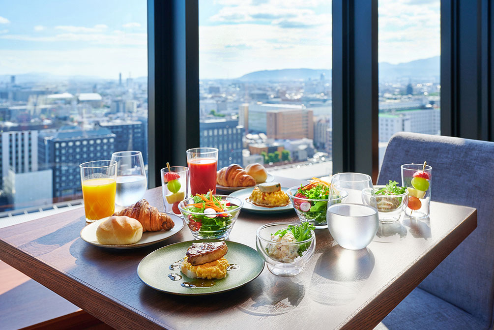 大きな窓から見える高層階からの景色と、パンやサラダ、お肉やフルーツなど和洋様々なお料理とドリンクがテーブル席に並べられている