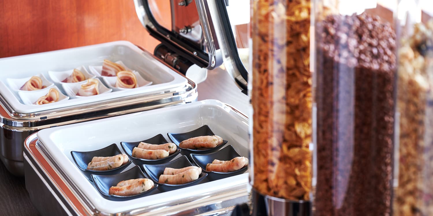 保温容器に入ったソーセージとベーコン、そして手前にはシリアルディスペンサーが並ぶビュッフェ形式の朝食。