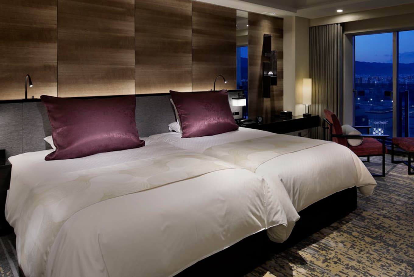 夜景が見える窓際の椅子、木目調の壁が特徴の、ツインベッドが置かれた高級感のあるホテルの客室。