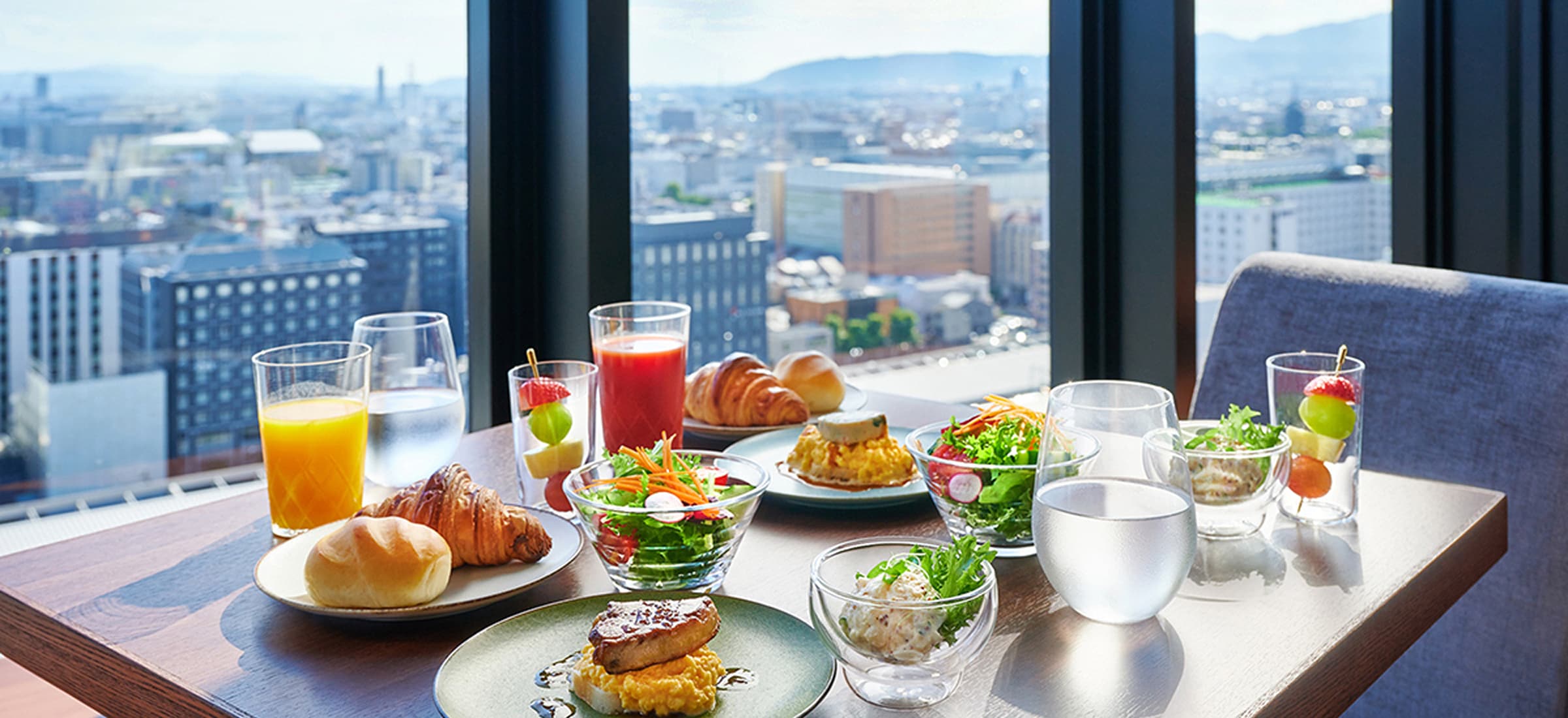 街を見下ろす窓際で撮影された、美味しそうな朝食（パン、サラダ、卵料理など）。
