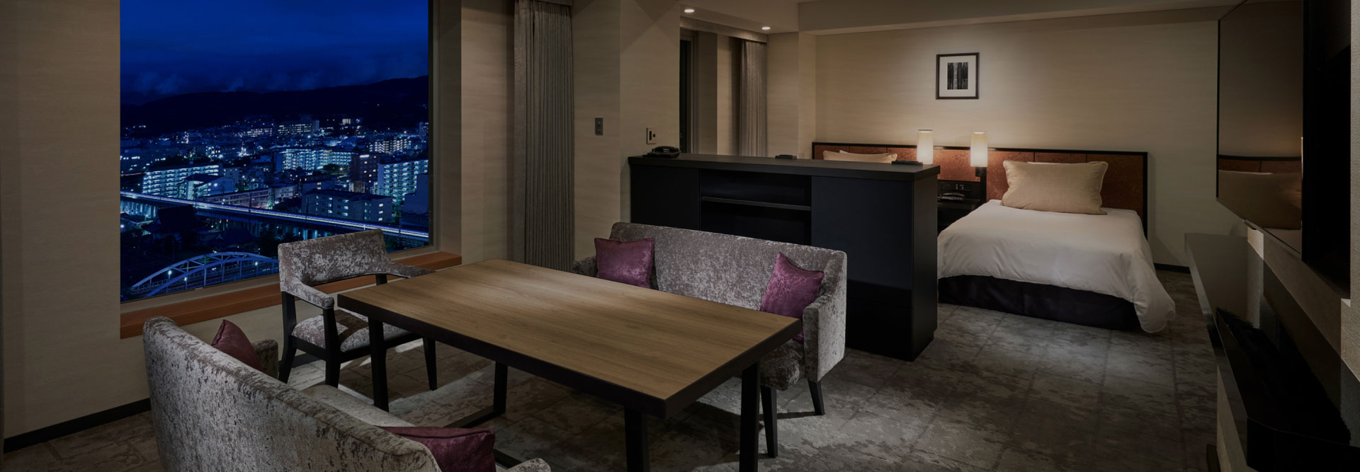 夜景を望む窓際にダイニングテーブルとソファを配置した、ベッドルーム一体型のシックな客室